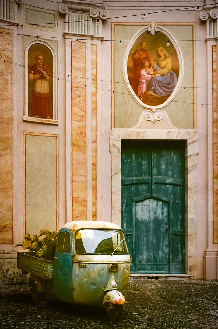 Eine kirchlich bemalte Ape vor dem Portal einer italienischen Kirche eines kleines Bergdorfs
