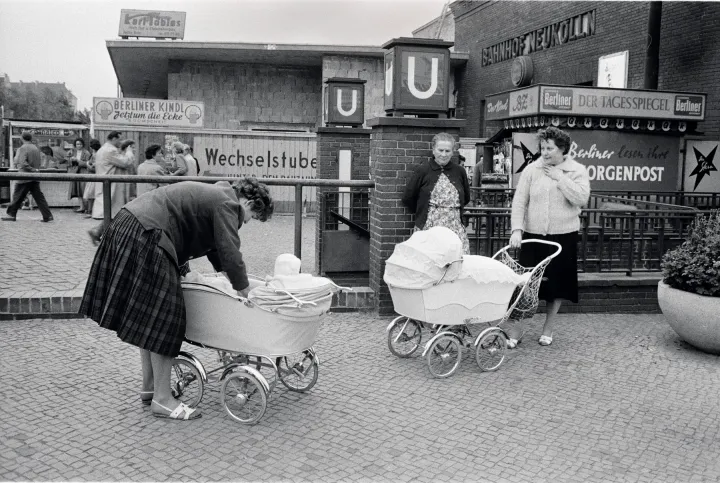 Schwarzweißfotografie eines belebten Platzes vor dem U-Bahnhof Neukölln in Berlin. Im Vordergrund beugt sich eine Frau über einen Kinderwagen, in dem ein Baby sitzt. Daneben stehen zwei weitere Frauen neben einem zweiten Kinderwagen. Im Hintergrund sind der Eingang zur U-Bahn mit großen „U“-Schildern, ein Zeitungskiosk sowie mehrere Passanten zu sehen. Die Szene vermittelt einen Eindruck des städtischen Alltags in Berlin der 1960er-Jahre.