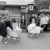 Schwarzweißfotografie eines belebten Platzes vor dem U-Bahnhof Neukölln in Berlin. Im Vordergrund beugt sich eine Frau über einen Kinderwagen, in dem ein Baby sitzt. Daneben stehen zwei weitere Frauen neben einem zweiten Kinderwagen. Im Hintergrund sind der Eingang zur U-Bahn mit großen „U“-Schildern, ein Zeitungskiosk sowie mehrere Passanten zu sehen. Die Szene vermittelt einen Eindruck des städtischen Alltags in Berlin der 1960er-Jahre.